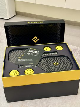 A Binance branded gift box (yellow and black) opened to reveal skateboard-like accessories and merchandise items with Binance logos and QR codes inside.