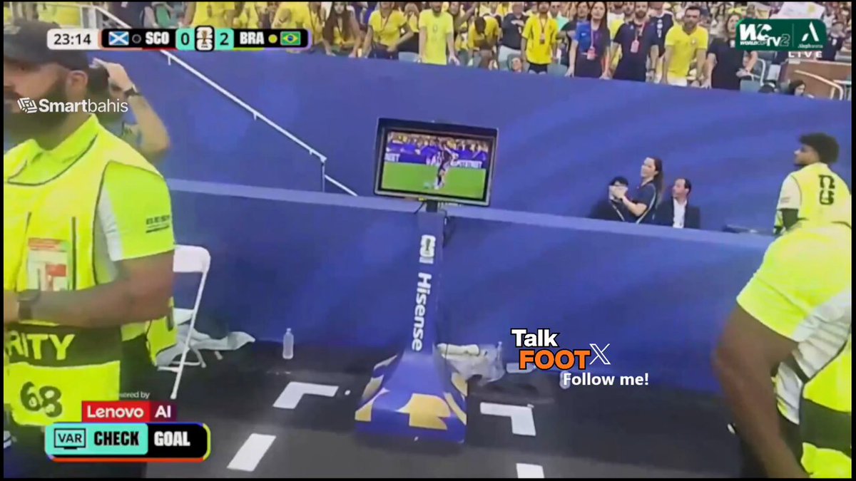 The image shows an electronic scoreboard with a soccer game happening, displaying some information about football goals and player names. The scoreboard also has text that encourages viewers to check out the online chatbot named Talkfoot for more details. In addition to the main screen, there is a small TV positioned in front of it. Several people are gathered around watching the game, with one person standing near the top edge of the image wearing an orange vest and holding a clipboard. Another person at a table stands closer to the center-left side of the image. The text on the scoreboard emphasizes the benefits of chatting with this bot for anyone interested in the game's details or scores.