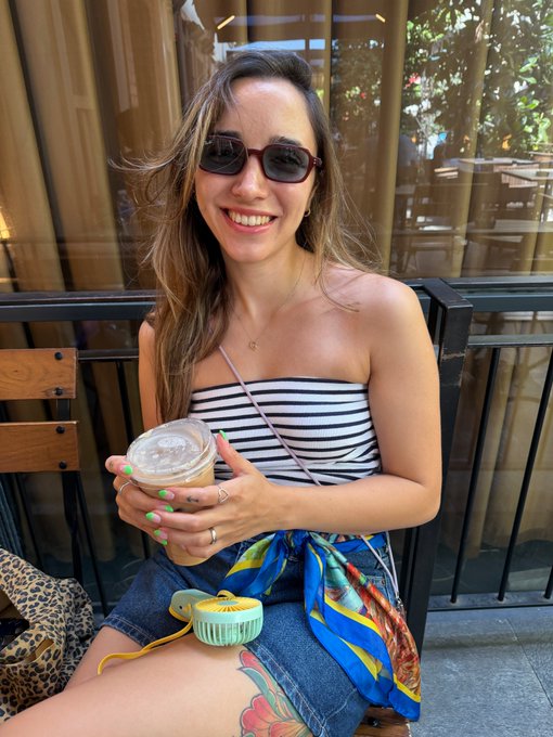 A smiling woman wearing sunglasses and a striped top sits outdoors holding an iced coffee, with a small portable fan resting on her lap.