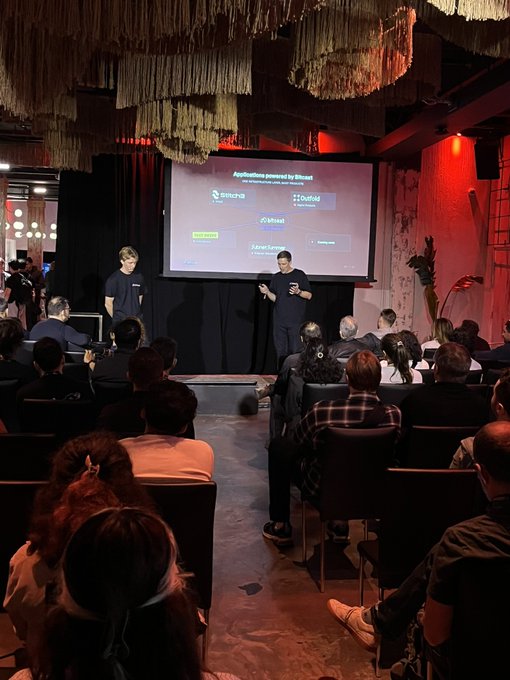 Two men stand on stage presenting in front of a screen displaying "Applications powered by Bitcast" with logos for Sencha, Oxfield, dSheel, and FluxNet, as they announce two new apps from Bitcast Network aimed at expanding Bittensor's reach and enhancing the creator economy to an audience seated in a dimly lit venue with hanging decorative fringes.