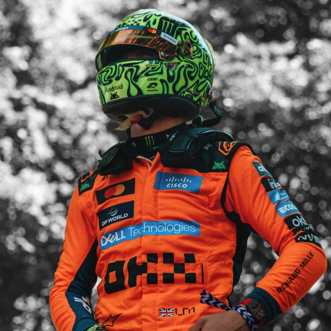 A Formula 1 driver in an orange McLaren racing suit and a green patterned helmet with Monster, Android, and Cisco logos stands focused, with visible sponsor patches including Dell Technologies, OKX, and Richard Mille—capturing the intense pre-race concentration reflected in the caption "Locked in to run it again."