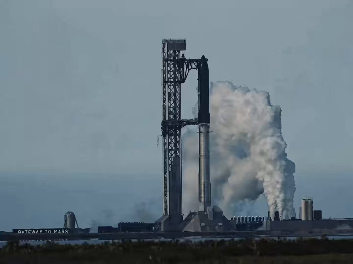 The image shows a large rocket launch site with a tall, gray structure that appears to be a space shuttle launchpad or a satellite launchpad. The sky above the launchpad has a hazy blue color, suggesting an overcast day. There is also some smoke coming from the launchpad, indicating that a rocket launch might have recently taken place.