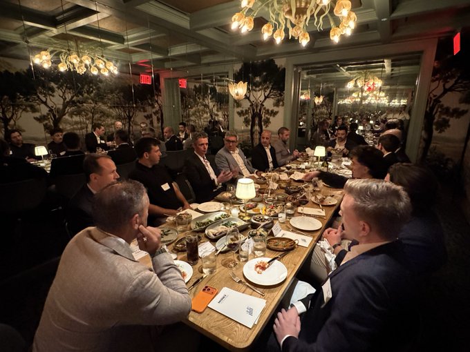 A group of people seated at a long table enjoy a meal together in an elegant restaurant with ornate chandeliers and decorative wallpaper.