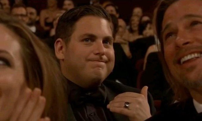 Jonah Hill, wearing a black tuxedo, smiles and gives a thumbs-up while seated in an audience at what appears to be an awards ceremony.