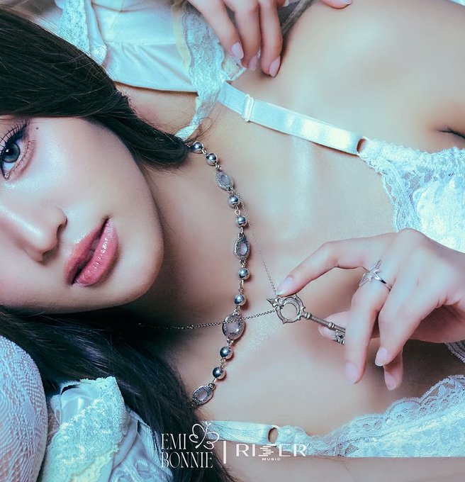 An artistic promotional image for EMI BONNIE x RISER MUSIC featuring a woman with dark hair wearing a silver jewelry-adorned harness over a white lace top, holding a decorative key.