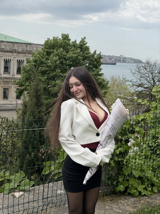 A young woman with long dark hair posing outdoors in a white blazer over a burgundy top and black skirt, holding a newspaper, with a waterfront view featuring a historic building and ships in the background.