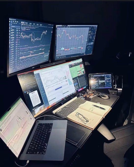 A multi-monitor trading setup in a dark room displaying financial charts and data, alongside a laptop, notebook with sketches, glasses, and a pen on a desk.