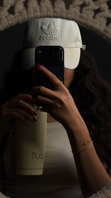 A person wearing an Adidas cap takes a mirror selfie while holding a phone and a tuff drink, their face obscured.