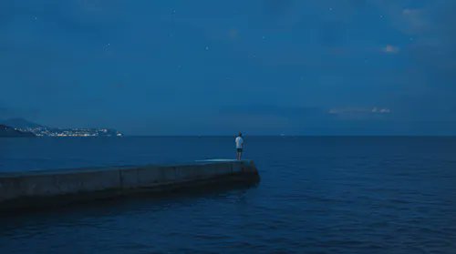 A nighttime scene shows a person standing at the end of a pier extending into a dark blue sea under a starry sky, with distant city lights visible on the horizon.