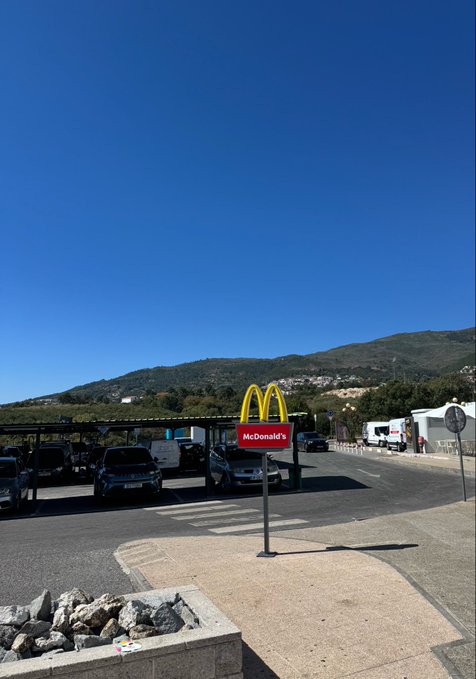 A McDonald's restaurant with its iconic golden arches sign stands against a backdrop of rolling green hills under a clear blue sky, with several cars parked under the canopy. The image relates to "GM bullas" as it appears to be taken in Bullas, a town in the Murcia region of Spain, showcasing a local McDonald's location in this scenic rural setting.