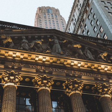 The image shows the grand facade of the New York Stock Exchange (NYSE), with its iconic columns, ornate gold detailing, and the inscription "NEW YORK STOCK EXCHANGE" clearly visible, alongside a towering skyscraper in the background. This historic landmark ties directly to the announcement, as NYSE's parent company ICE is partnering with OKX to enable onchain trading of 63 tokenized U.S. stocks under the SEC's innovation exemption.