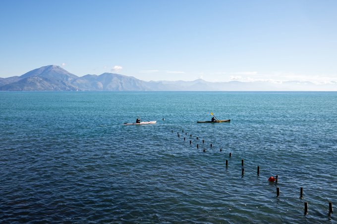 Two kayakers paddle on a calm blue sea with mountains rising in the distant background, near a line of wooden posts extending into the water.