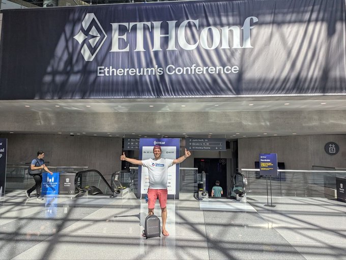 The image features a man standing next to an escalator, holding his arms outstretched as if he's about to give a speech or make a gesture. The man appears to be wearing a white shirt and red shorts. In front of him is a large banner that says "ETHConf" in black letters. There are also two other people visible in the image - one person standing near the escalator, and another person walking away from it.