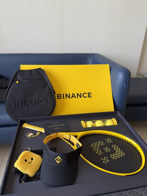 An open Binance branded gift box displays various yellow and black promotional merchandise including a tennis racket, cap, bag, and plush toy arranged in foam inserts.