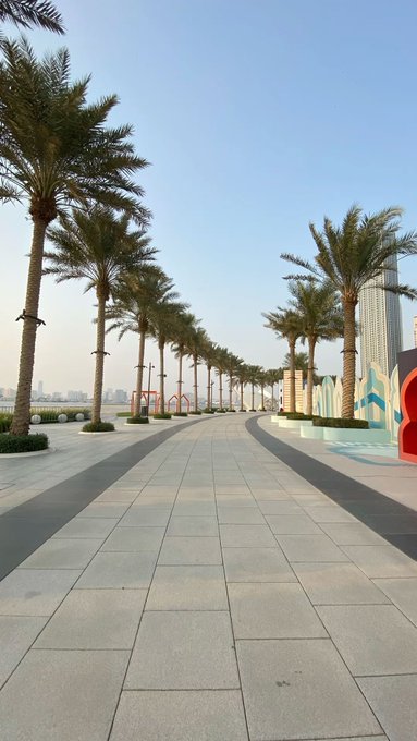 A wide, sunlit promenade lined with tall palm trees leads toward a modern waterfront, where a tall skyscraper is visible on the right and colorful geometric wall art decorates the adjacent plaza. The serene, upscale setting evokes the "GM" lifestyle of a Grand Morning stroll through a luxurious coastal city.