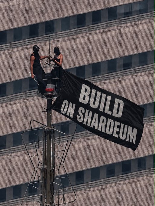Two masked individuals unfurl a large black banner reading "BUILD ON SHARDEUM" from atop a communications tower or antenna structure, set against a grid-patterned building facade—capturing a public demonstration that visually reinforces the accompanying message.