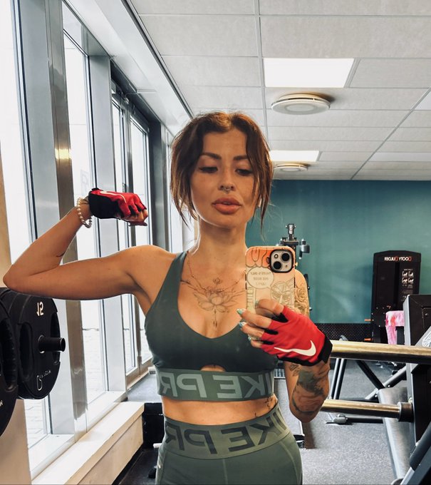 A woman in a green Nike Pro sports bra and leggings takes a mirror selfie in a gym while flexing her arm, wearing red Nike workout gloves, with a "12" kg weight plate visible behind her—capturing the determined mindset of being "1% better than yesterday."