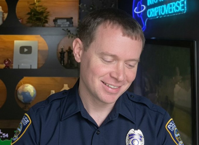 A smiling police officer in a dark uniform with a badge and shoulder patches sits in front of shelves displaying a YouTube play button, a globe, and a 'CRIPTOVERSE' neon sign.