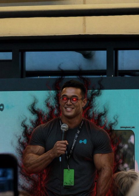 A muscular man wearing red-tinted sunglasses and a dark t-shirt smiles while holding a microphone in front of a teal backdrop with red flame-like graphics and the text '@theghost_ai' visible.