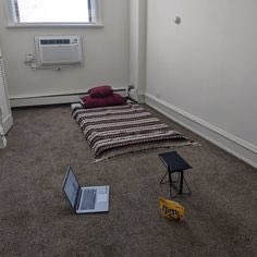 An overhead view of a sparse room with carpeted flooring, featuring a striped blanket with a pillow, a small folding table, a "CAUTION" wet floor sign, and an open laptop on the floor.