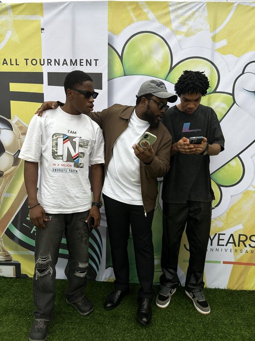 Three young men pose in front of a green and white "BALL TOURNAMENT" and "YEARS ANNIVERSARY" backdrop, with the two on the right intently checking their phones while the third stands in sunglasses and a "TAM ONE" graphic tee, humorously illustrating the joke about chasing a high-value crypto tip that could "print $50k."
