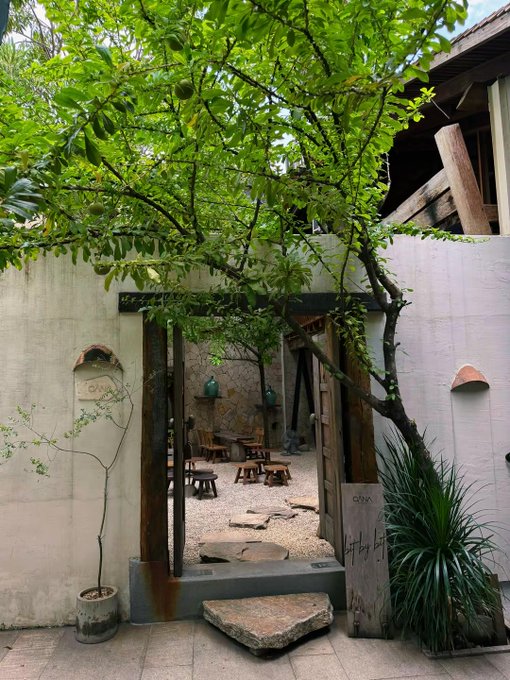 A view through a doorway into a courtyard with stone walls, surrounded by green foliage and tree branches, with a sign reading 'OAK' visible.