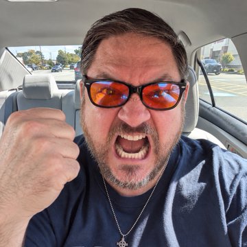 A bearded man wearing reflective orange sunglasses and a black shirt, shouting angrily with a clenched fist while sitting inside a car.