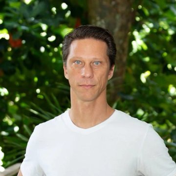 A man with short brown hair and blue eyes poses outdoors against green foliage, wearing a plain white t-shirt.