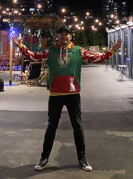 A person poses with arms outstretched wearing a green, red, and yellow hoodie featuring a white bunny character, standing in a lit outdoor plaza at night. The hoodie's design appears to mirror the "Doodles" toy character referenced in the post.