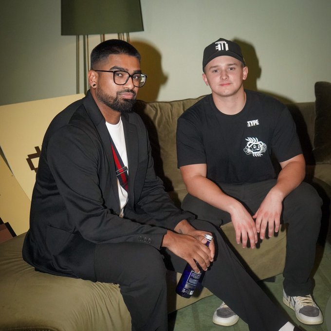 Two men sit on a beige couch indoors; the man on the left wears a black suit, glasses, and has a beard, while the man on the right wears a black 'TYPE' t-shirt, a black cap, and holds a blue beverage can.