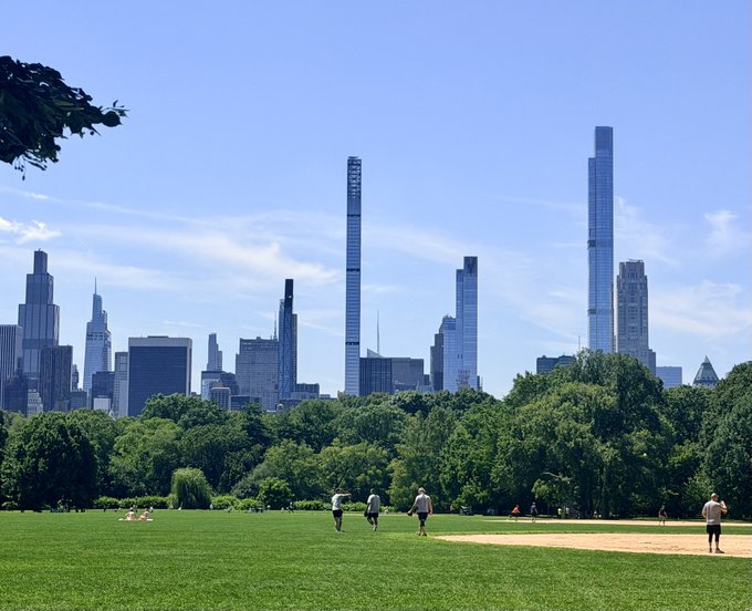 A group of people enjoy a casual baseball game on a lush green field in a park, with a stunning Chicago skyline featuring the prominent 432 N. Michigan Ave skyscrapers rising in the background under a clear blue sky — the perfect backdrop for watching investments soar to new heights.