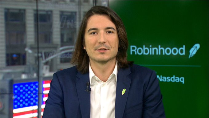 A man with long brown hair wearing a navy blazer and white shirt speaks in a studio setting, with a 'Robinhood' logo and 'Nasdaq' text visible on the screen behind him alongside an American flag.