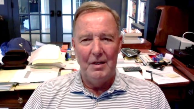 A man in a striped polo shirt sits in an office with a wooden desk covered in papers and office supplies, appearing to speak on a video call.