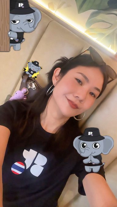 A young woman with long dark hair smiles for a selfie, wearing a black t-shirt and hoop earrings with cartoon elephant stickers from a social media filter overlaid on the image.