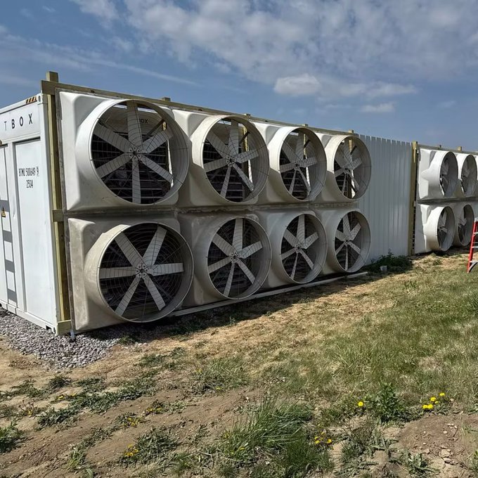A photograph of large industrial cooling fans mounted on white container-style units at an outdoor installation under a partly cloudy sky.