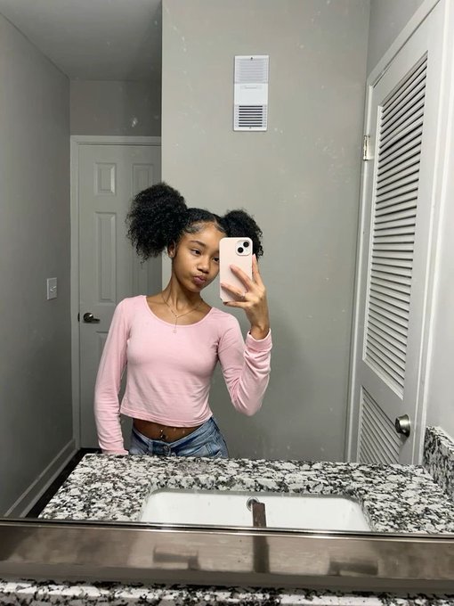 A mirror selfie of a young woman with curly hair styled in two puffs, wearing a pink long-sleeve crop top and jeans, standing in a bathroom with a granite countertop.