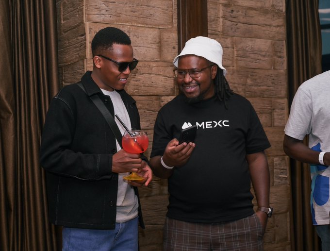 Two men pose together at an event, one holding a red cocktail while wearing a black jacket and sunglasses, and the other in a white bucket hat and glasses holding a phone, with a black t-shirt prominently displaying the white "MEXC" logo. The image visually supports the post by capturing the MEXC Africa Kenyan Ambassador and the Head of MEXC Growth in Africa networking at the Block and Chill event.