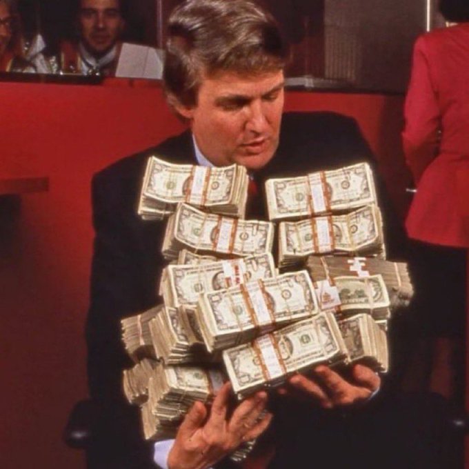 A man in a suit sits while attempting to hold an enormous, precariously stacked pile of bundled cash. The scene humorously visualizes the casual "Gm" (good morning) greeting as a playful exaggeration of overwhelming wealth or financial success to start the day.