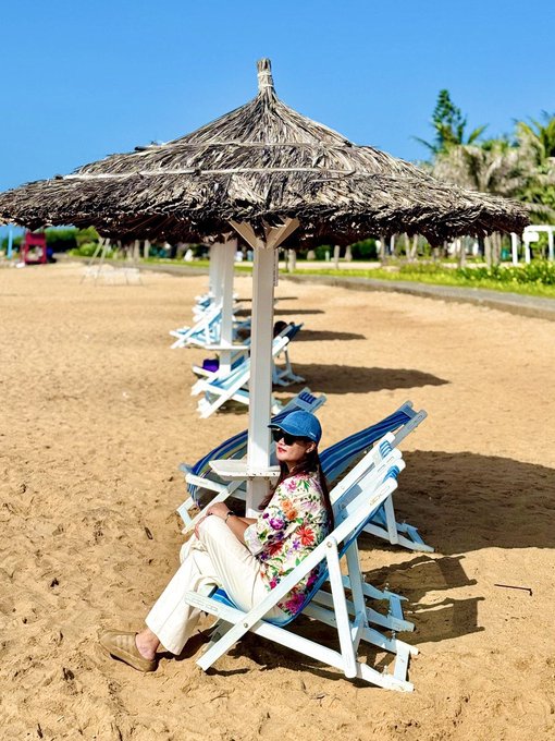 A woman in a floral shirt and cap relaxes under a thatched beach umbrella, embodying the "living very chill" lifestyle amid the contrasts of online trading culture.