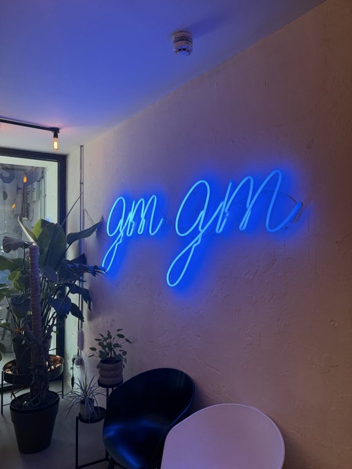 A blue neon sign on a wall reads 'gm gm' in cursive script, mounted above black and white chairs with potted plants nearby.