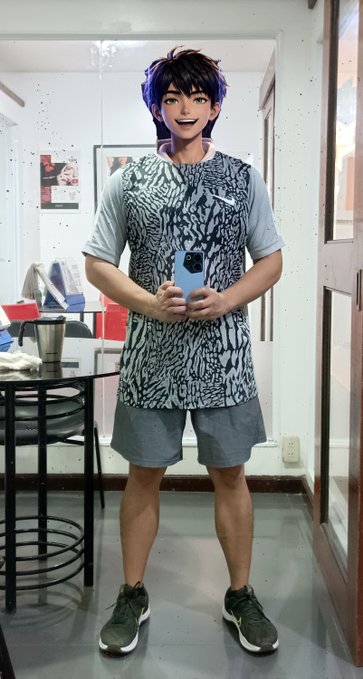 A man stands in an office hallway wearing a short, patterned tunic over a grey shirt and shorts, holding a blue smartphone with an animated cartoon face filter applied to his head.