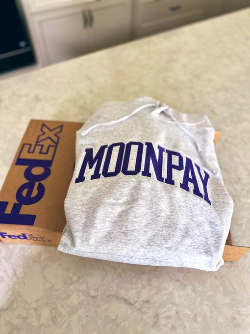 A folded gray tank top featuring "MOONPAY" in purple block lettering rests on top of a FedEx shipping envelope on a kitchen countertop, suggesting a recent delivery of MoonPay merchandise just in time for "hoodie szn."