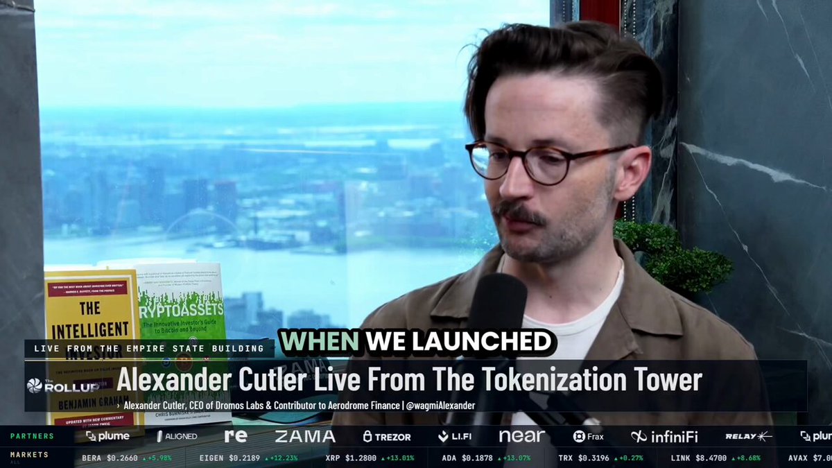 The image shows a man wearing glasses, standing next to a window with a view of a city skyline outside. The text on the screen reads "When We Launched", which suggests that there might be an event or launch related to cryptocurrency taking place at this location.