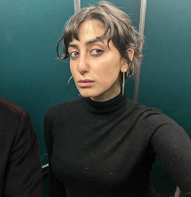 A selfie-style photo of a young woman with short dark hair, large hoop earrings, and a black turtleneck top taken in front of a green tiled wall.