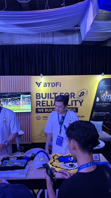 A BYDFi booth at a crypto conference features a representative in a branded t-shirt displaying the slogan "BUILT FOR RELIABILITY" and "2020 Founded" alongside merchandise, while a young attendee in the foreground browses items on the table. The image captures the exchange's promotional presence at the event, aligning with @BYDFi's social media showcasing their industry engagements.