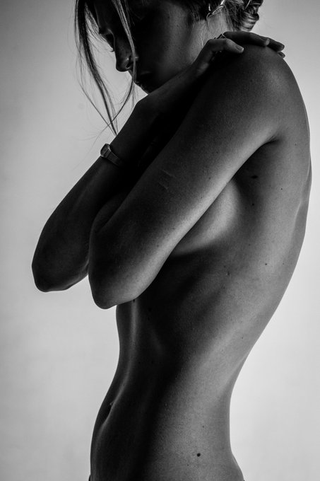 A black and white photograph showing the back and side profile of a nude person with long hair, arms crossed over their shoulders.