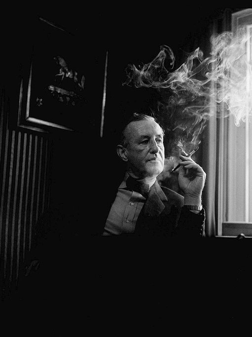 A black and white photograph of an older gentleman in a suit smoking a cigarette, with smoke curling up into the air in a dimly lit room.