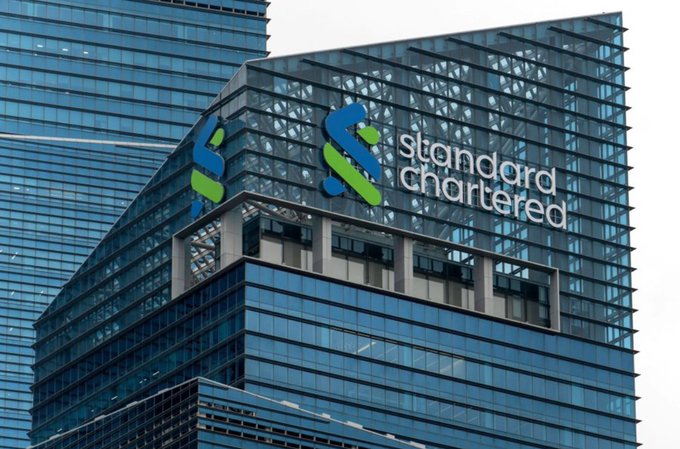 The Standard Chartered logo is displayed on a modern glass skyscraper. The image relates to the post by showcasing the bank's involvement in launching the regulated $HKDAP Hong Kong dollar stablecoin through its Anchorpoint initiative, highlighting Standard Chartered's role in advancing institutional digital finance infrastructure.