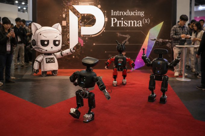 The image features three robots walking across a red carpet at an event. The robots are dressed in black outfits with white accents, giving them a futuristic appearance. They appear to be interacting with each other or with people around them. In front of the robots, there is a large poster that says "Introducing Prisma X". This poster likely provides information about the event and the company behind it. The background shows several people standing near the robots, observing their movements and interactions.