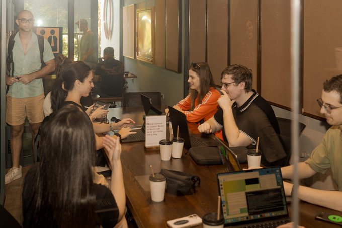 A group of people gathered around a wooden table in a cafe or co-working space, working on laptops and conversing.